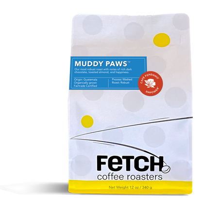 White Kraft paper coffee bag with a yellow band on the bottom and the Fetch logo with a bean next to the 'h'. The Muddy Paws label is a rich blue and there is a red Cafe Feminino sticker next to the Muddy Paws label.